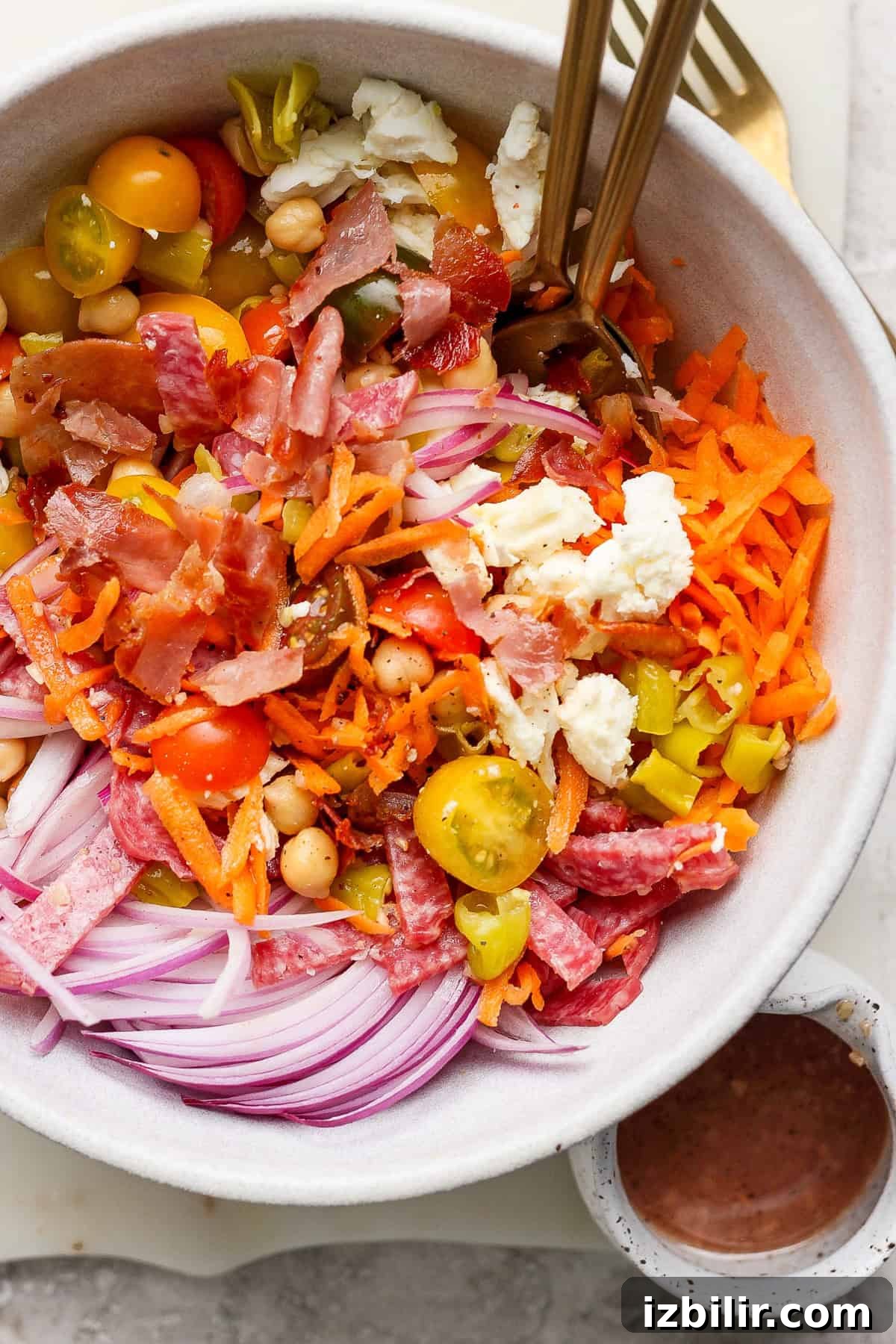 A bowl of salad with sliced red onions, cherry tomatoes, chickpeas, shredded carrots, crumbled cheese, and cured meats. Two forks are placed in the bowl. A small bowl of dressing is beside it.