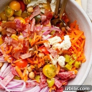 A salad bowl with shredded carrots, sliced red onions, cherry tomatoes, chickpeas, feta cheese, chopped bacon, and salami. Two forks are in the bowl.
