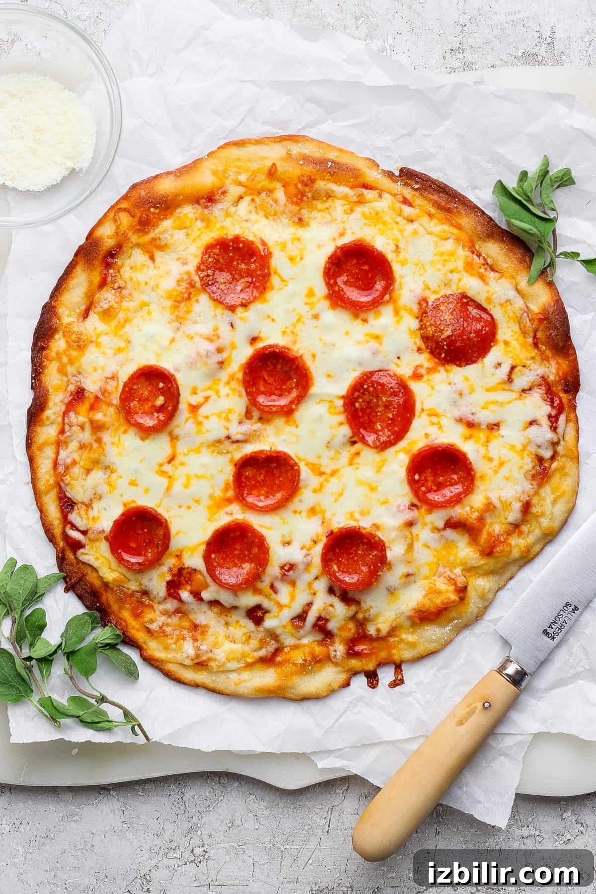 A pepperoni pizza on parchment paper with a knife beside it, garnished with herbs. A small bowl of grated cheese is in the top-left corner.