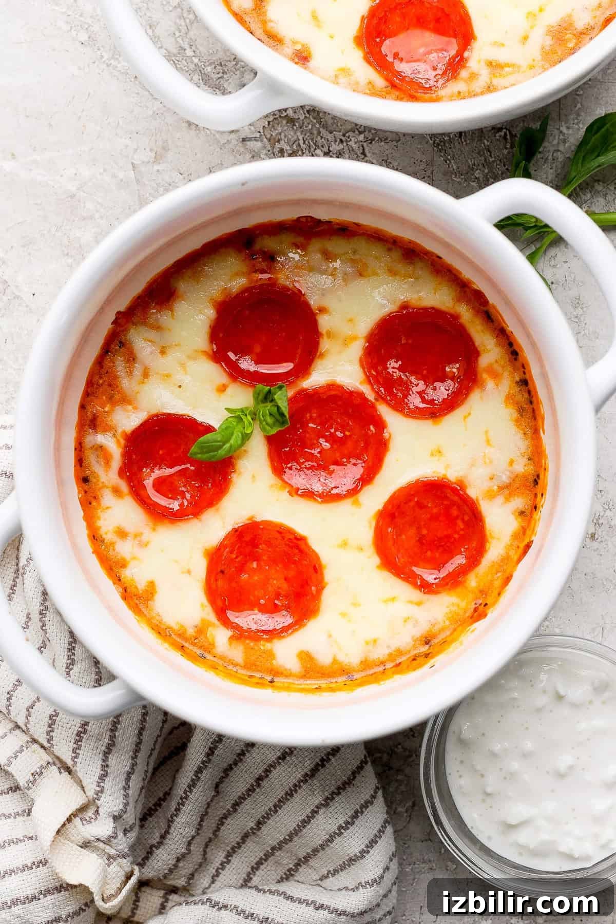 Single-Serve Cottage Cheese Pizza Bowl Recipe 2 A white bowl filled with baked pepperoni and cheese dip, garnished with a basil leaf, placed on a cloth next to a small bowl of dipping sauce.