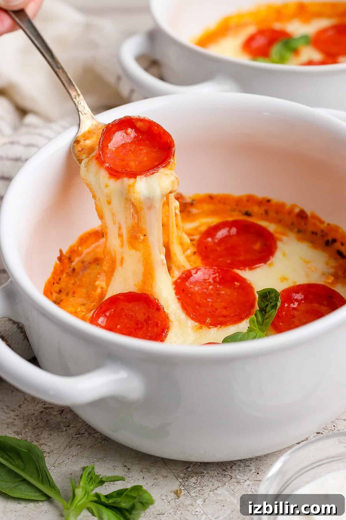 Single-Serve Cottage Cheese Pizza Bowl Recipe 4 A spoon lifts cheesy pepperoni pizza dip from a white bowl. Fresh basil leaves are visible nearby.
