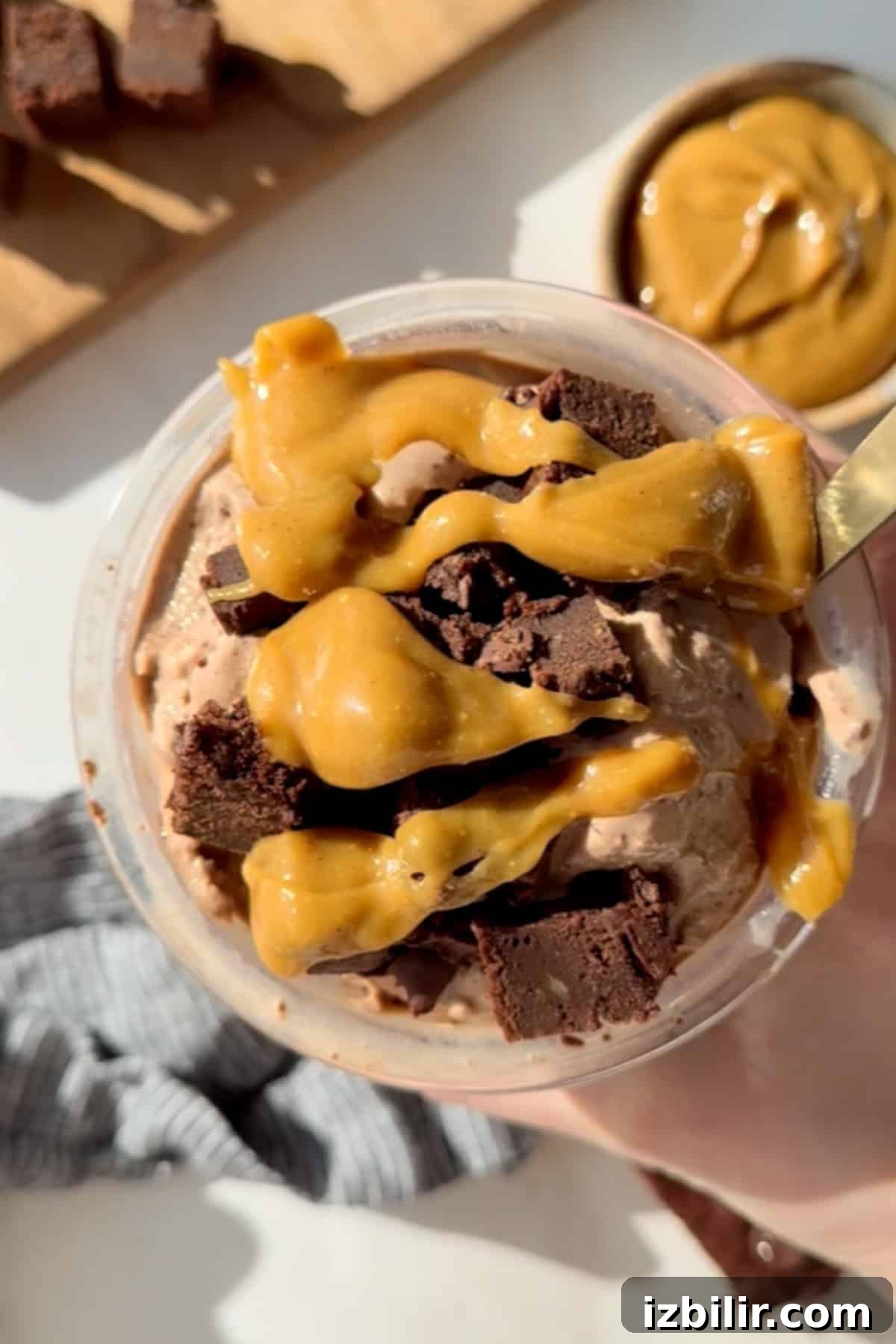 A cup of chocolate ice cream topped with caramel sauce and brownie chunks, with a spoon in it. A bowl with caramel is visible in the background.