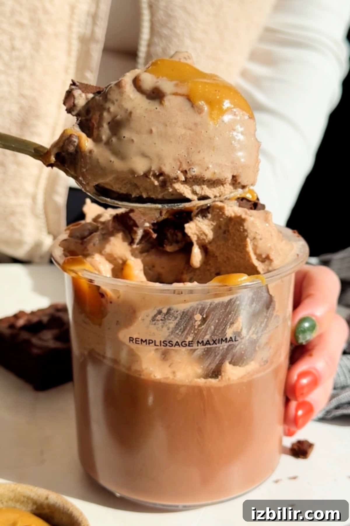 A person holds a spoon with ice cream drizzled with caramel over a clear cup, which seems filled to the maximum line. A brownie is visible in the background.