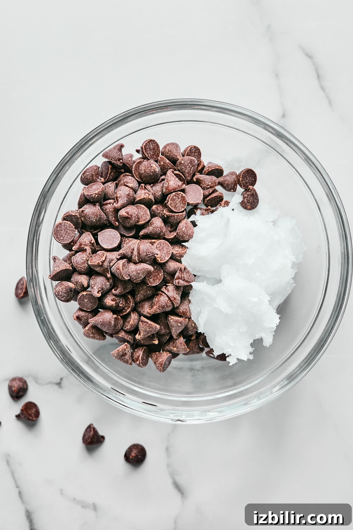 A glass bowl filled with chocolate chips and a dollop of coconut oil on a marble surface.