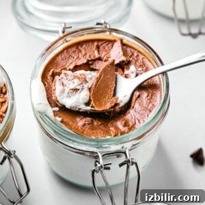 A spoon scoops chocolate dessert from a glass jar, revealing a creamy white layer underneath. Another jar with a similar dessert is partially visible in the background.