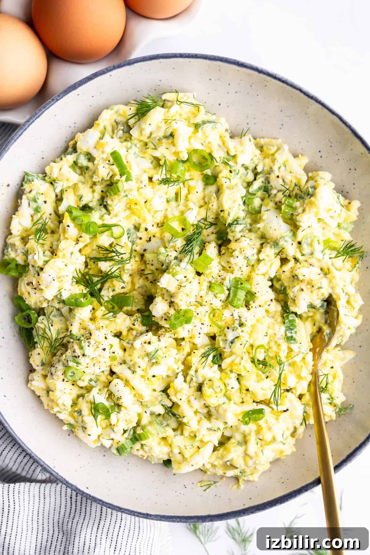 Creamy Cottage Cheese Egg Salad Recipe 2 A bowl of egg salad with chopped green onions and dill, seasoned with black pepper. Three whole eggs are in the background.