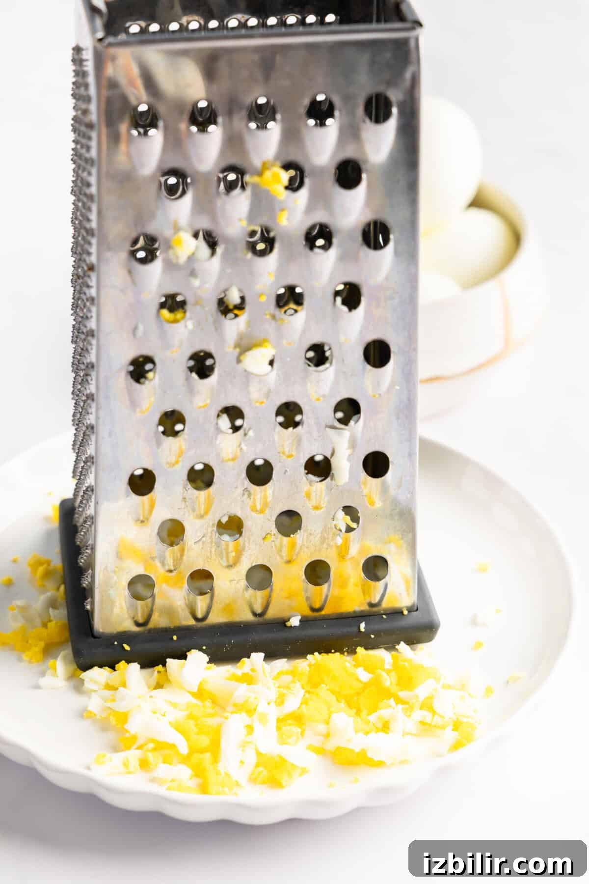 Creamy Cottage Cheese Egg Salad Recipe 3 A metal cheese grater on a white plate with finely grated egg, alongside a bowl containing whole boiled eggs.