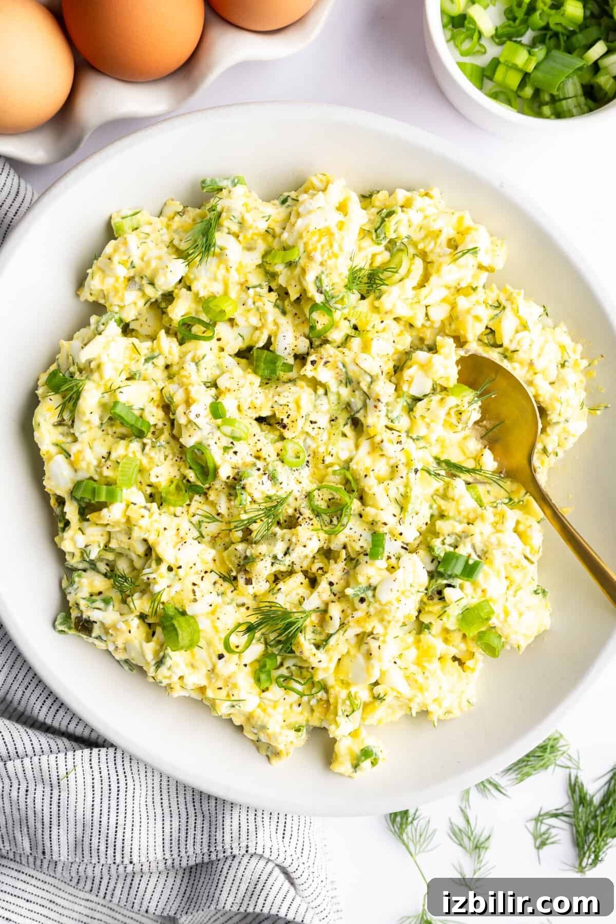 Creamy Cottage Cheese Egg Salad Recipe 4 A bowl of egg salad garnished with chopped green onions and dill, placed next to whole eggs, sliced green onions, and a striped cloth.
