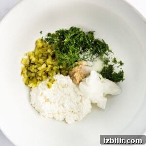 Creamy Cottage Cheese Egg Salad Recipe 5 White bowl containing cottage cheese, chopped pickles, fresh dill, minced garlic, salt, and sour cream.