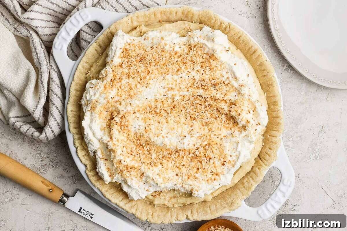 High-Protein Coconut Cream Pie Recipe 2 A pie topped with whipped cream and toasted coconut flakes, on a light-colored surface with a knife and striped cloth nearby.