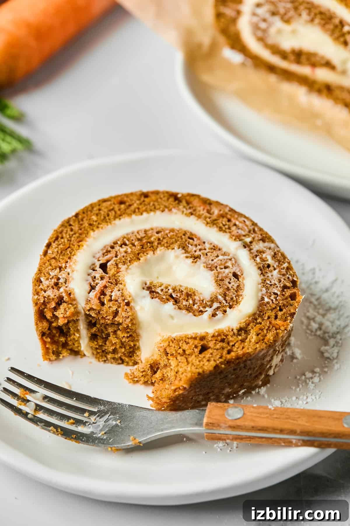 A slice of carrot cake roll with cream cheese filling on a white plate, beside a fork. A part of the roll is sliced off and a carrot is in the background.