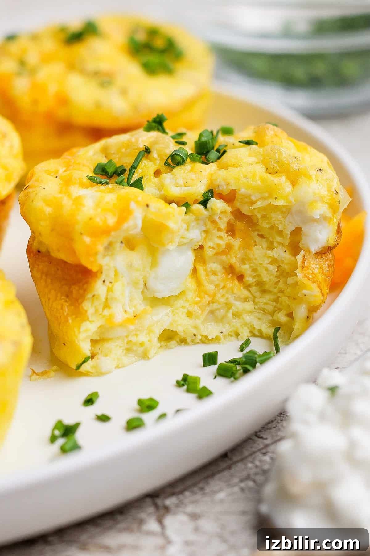 Creamy Cottage Cheese Baked Eggs Recipe 2 Close-up of a fluffy egg muffin topped with chopped chives, showing a bite taken out, on a white plate with blurred background.