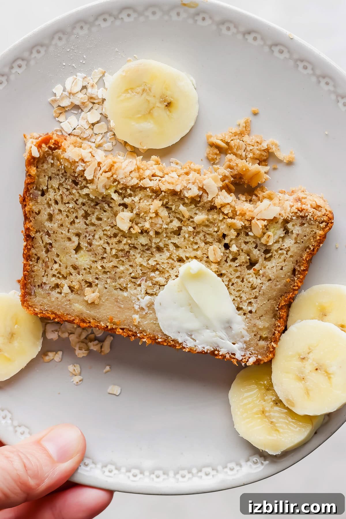 Slice of banana bread with butter and oats on a white plate, garnished with banana slices.