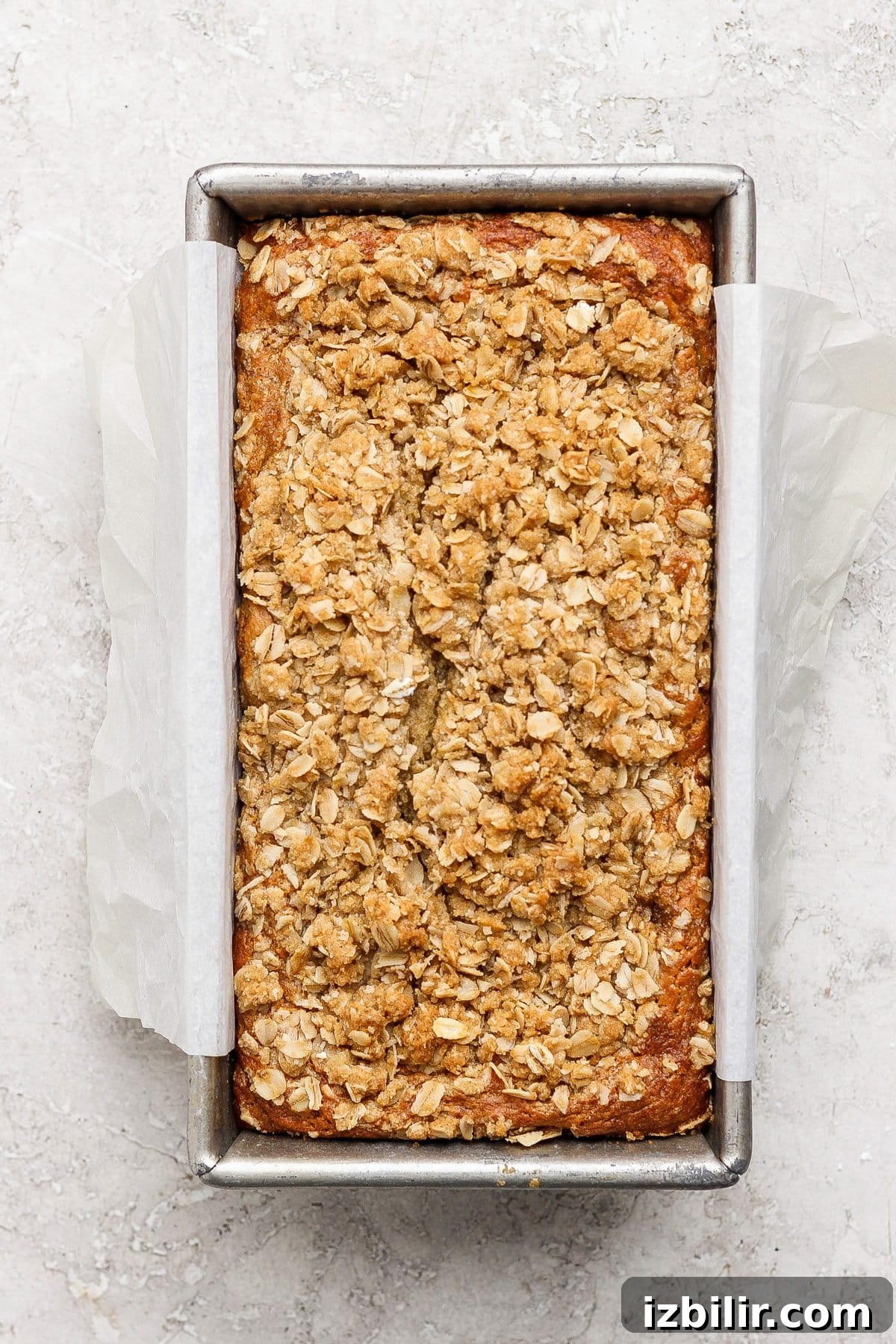 Loaf of banana bread with oat crumble topping in a parchment-lined metal baking pan on a light textured surface.