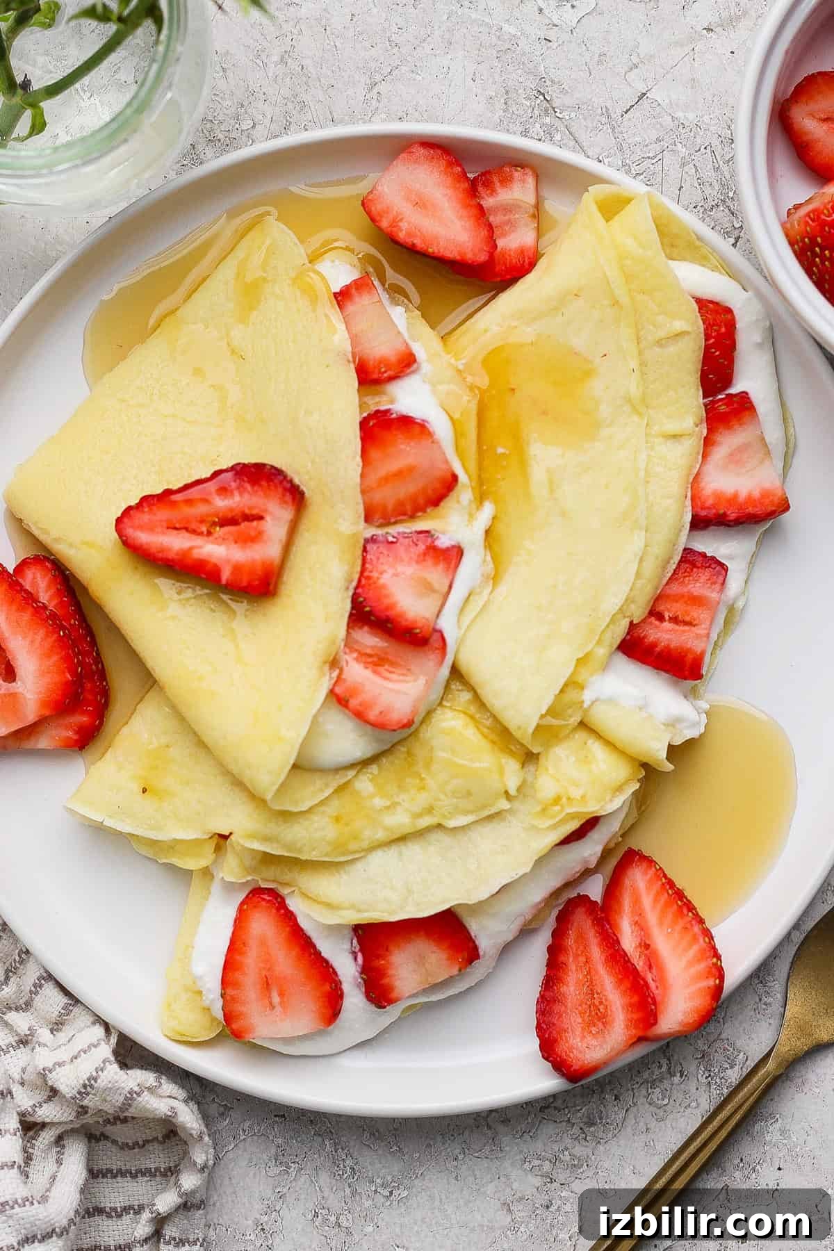 Crepes filled with cream and topped with sliced strawberries and syrup on a white plate. A fork and a striped napkin are beside the plate.