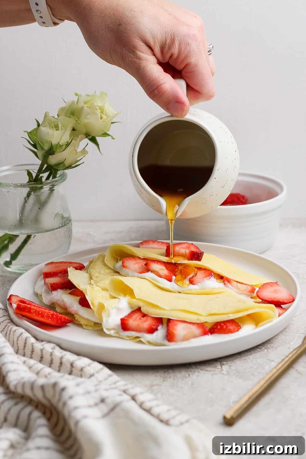 A hand pours syrup over crepes filled with strawberries and cream, placed on a white plate. A small vase with white roses and a striped cloth are in the background.