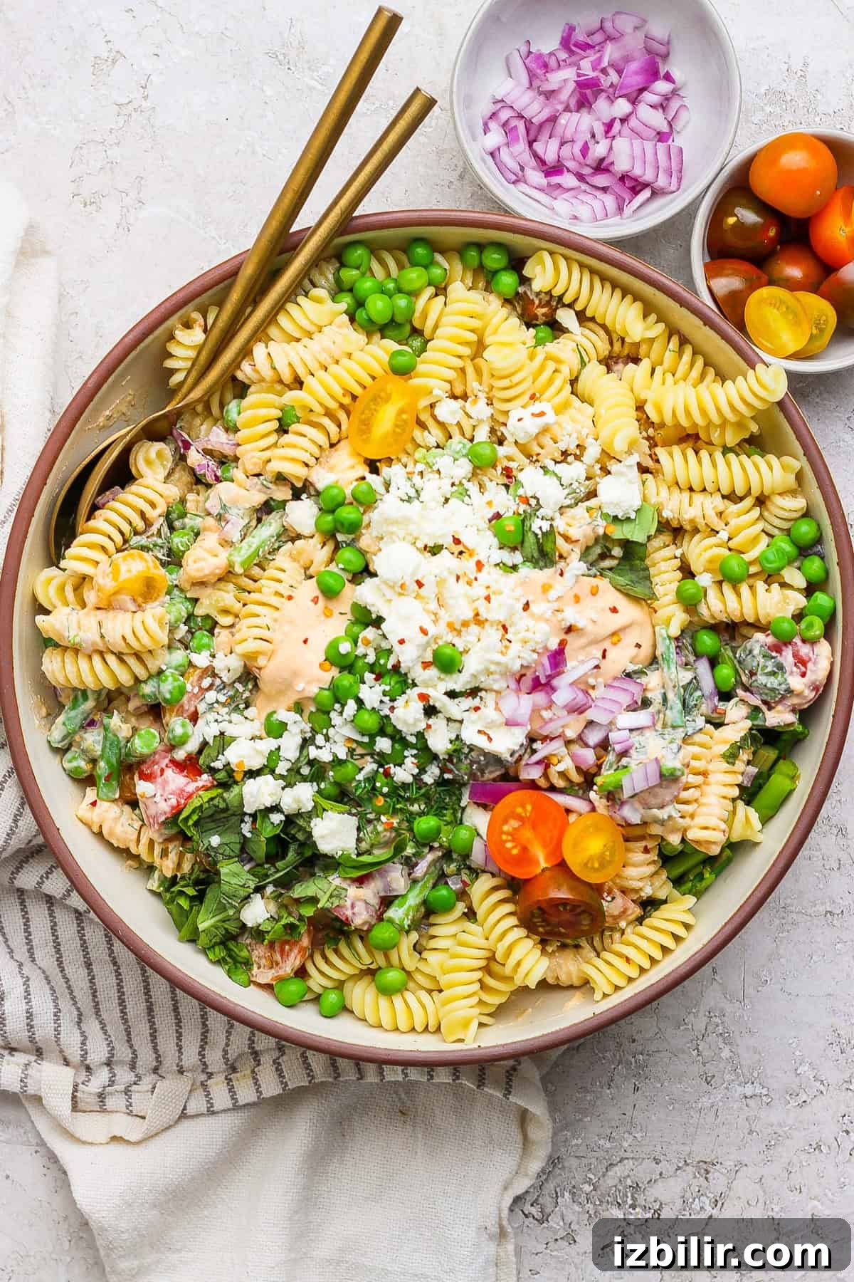 Roasted Red Pepper Pasta Salad with Cottage Cheese 2 A bowl of pasta salad with fusilli, cherry tomatoes, peas, feta cheese, and red onion. Two gold utensils rest on the side. Small bowls of red onions and cherry tomatoes are nearby.