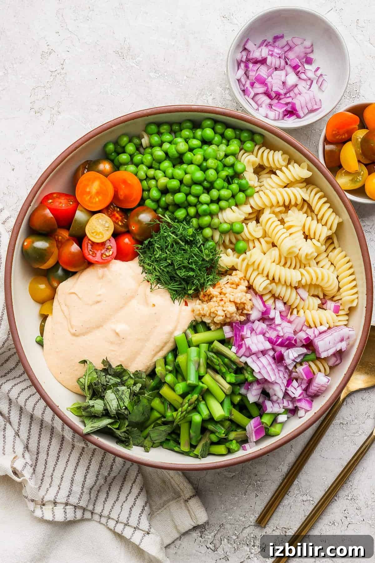 Roasted Red Pepper Pasta Salad with Cottage Cheese 5 A bowl with rotini pasta, cherry tomatoes, peas, chopped red onion, green beans, fresh herbs, and a creamy sauce. A small bowl of onions and cutlery are beside it.