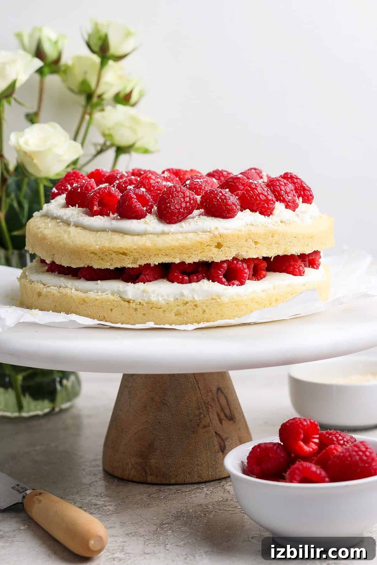 Two-layer vanilla cake with white frosting and raspberries on top, displayed on a wooden cake stand. White flowers and a bowl of raspberries are in the background.