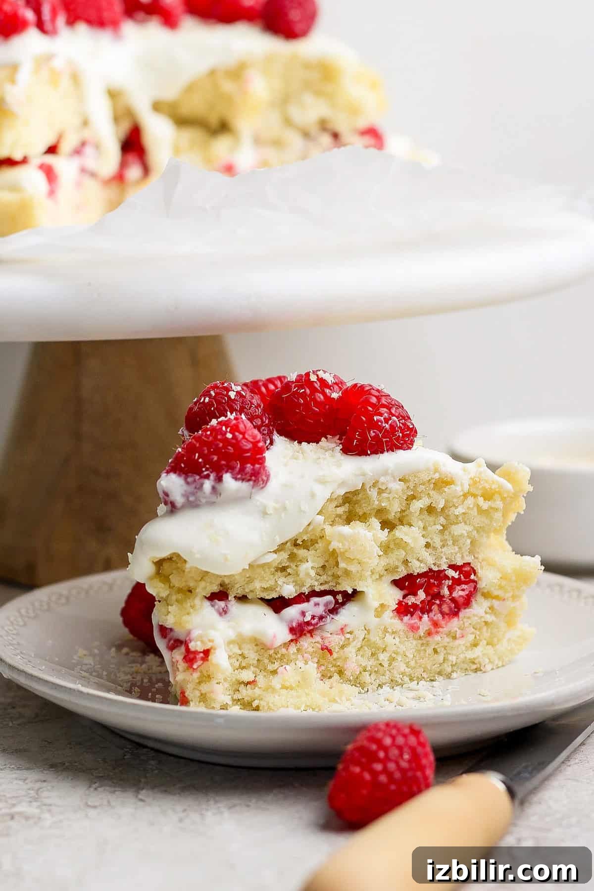 Slice of vanilla cake with white frosting and raspberries on top and inside, displayed on a plate with a wooden-handled knife beside it.
