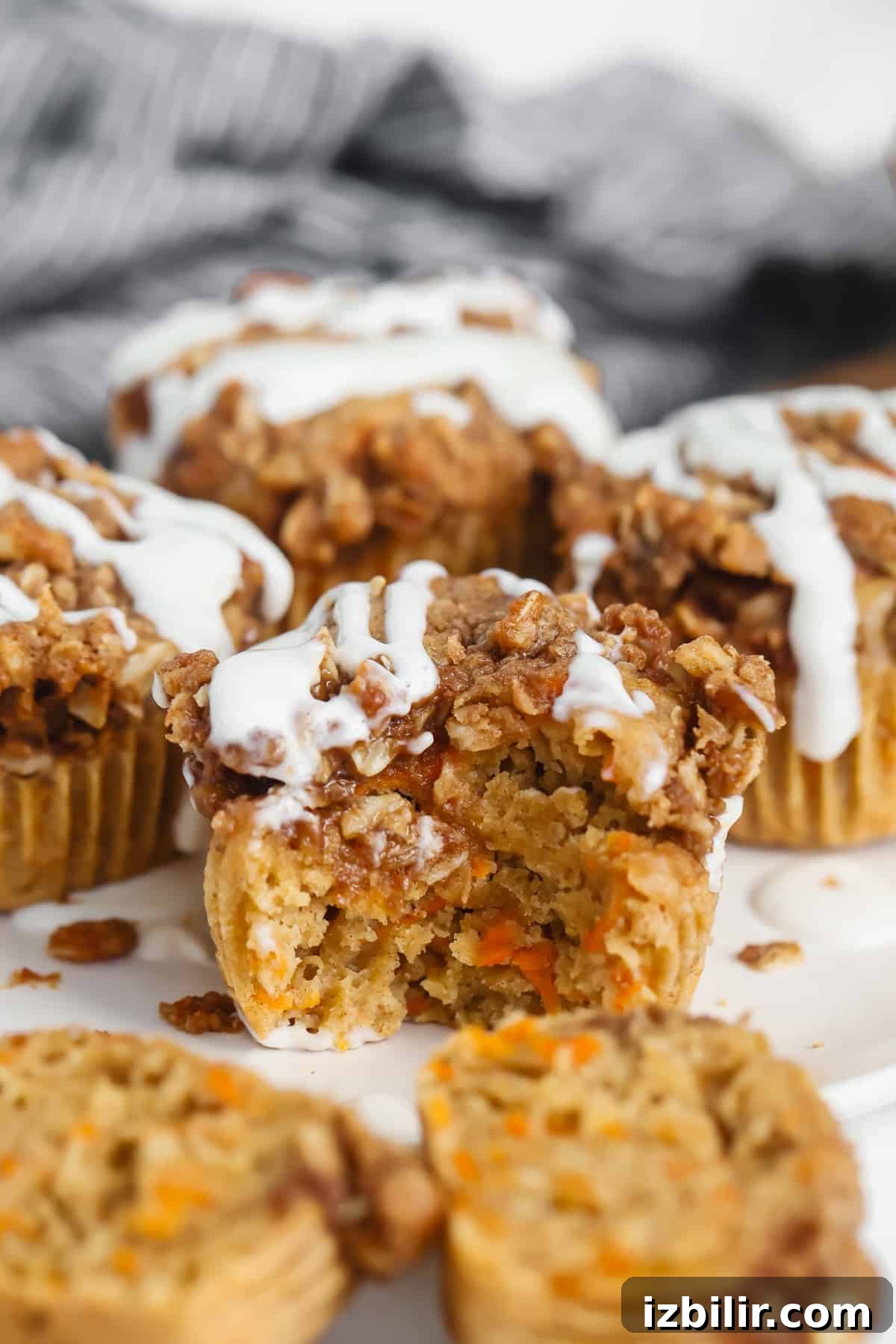 Carrot muffins with crumble topping and white icing drizzled on top, one muffin partially eaten, displayed on a white surface.