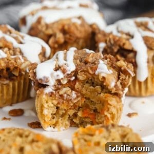 Close-up of a carrot muffin with a crumbly topping and white icing. One muffin has a bite taken out, revealing a moist interior with visible carrot pieces.