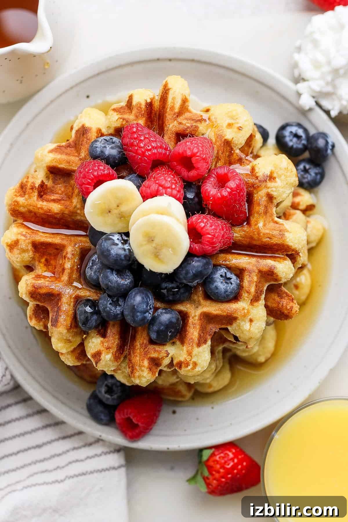 Fluffy Cottage Cheese Waffles Recipe for Brunch 2 A stack of waffles topped with blueberries, raspberries, and banana slices on a plate. A glass of orange juice and a striped napkin are nearby.