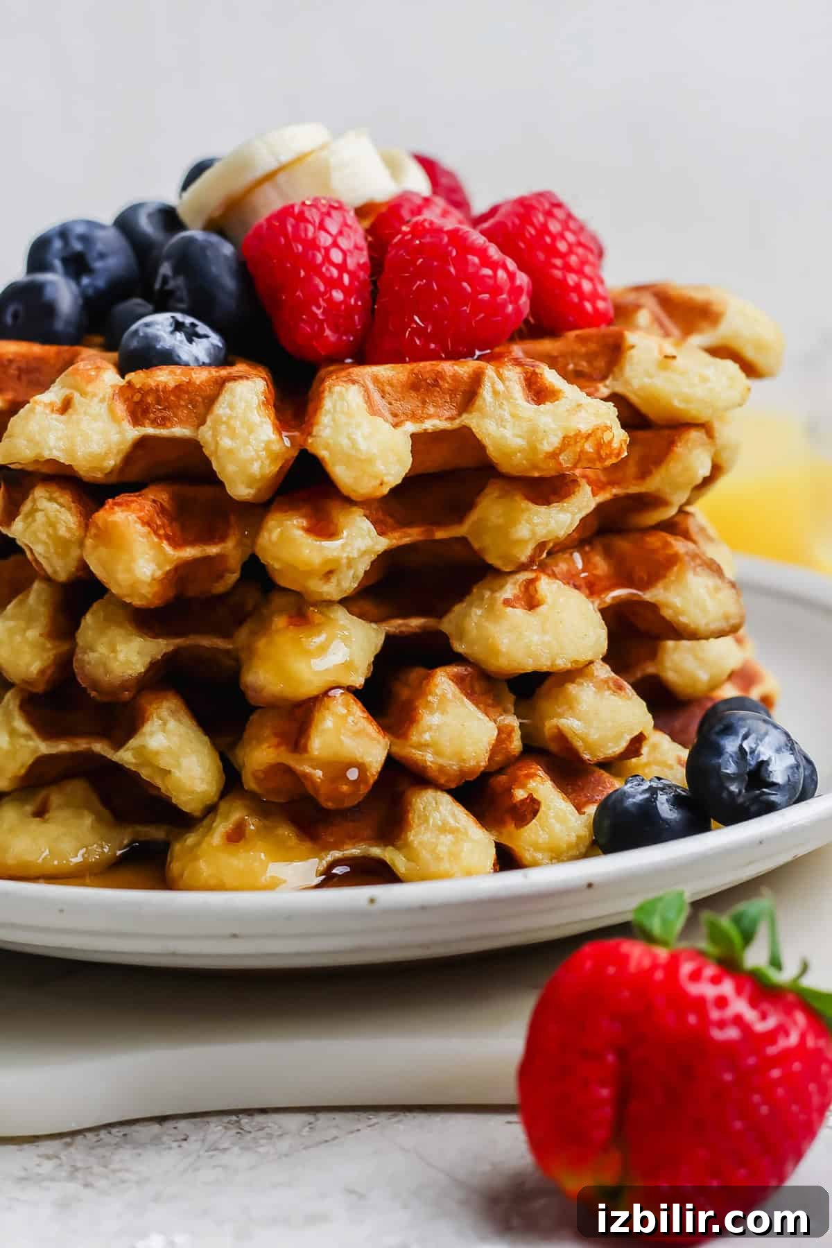 Fluffy Cottage Cheese Waffles Recipe for Brunch 5 A stack of waffles topped with banana slices, raspberries, and blueberries, drizzled with syrup. A strawberry sits on the plate's edge.