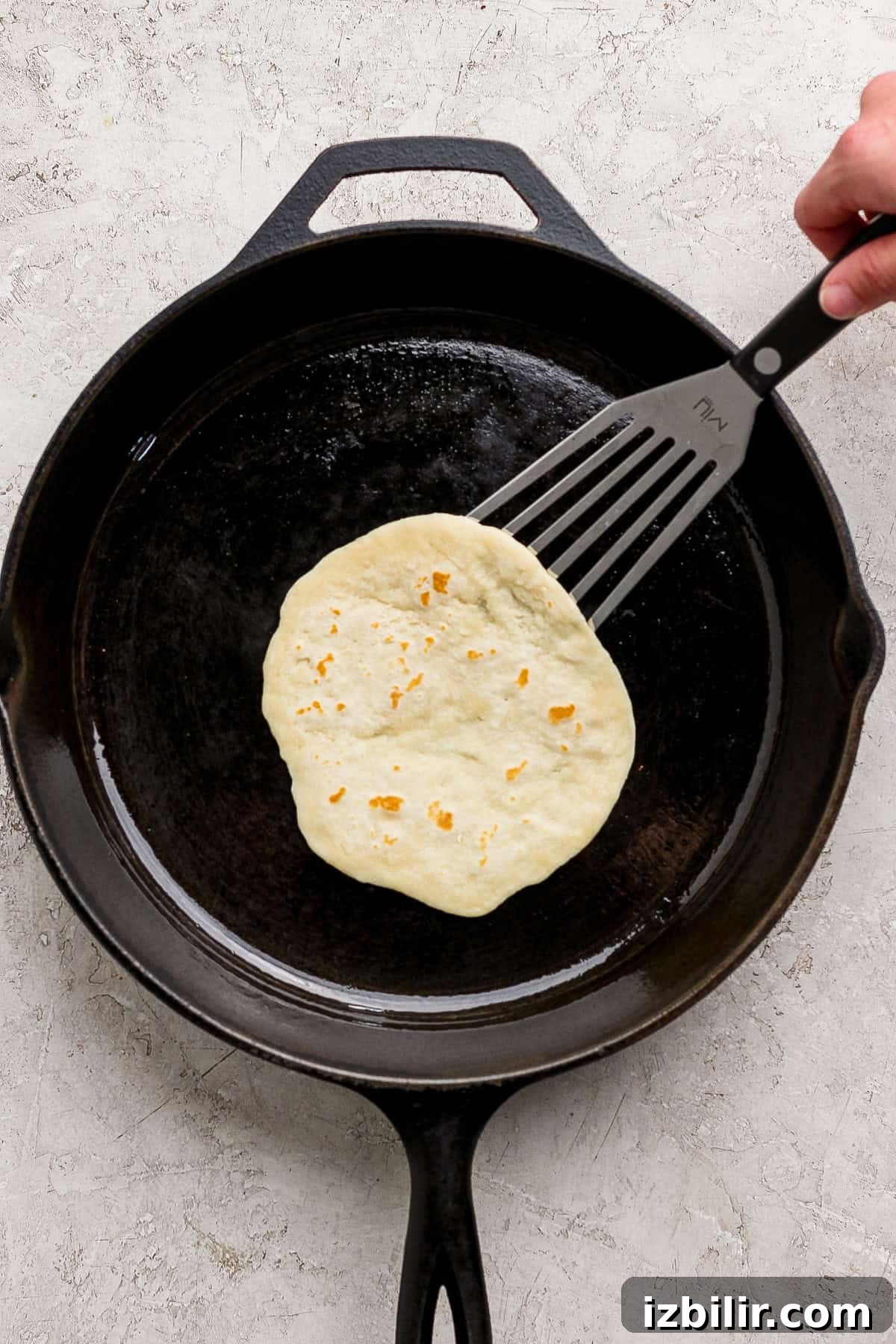 A hand uses a spatula to lift a tortilla cooking in a black cast iron skillet.
