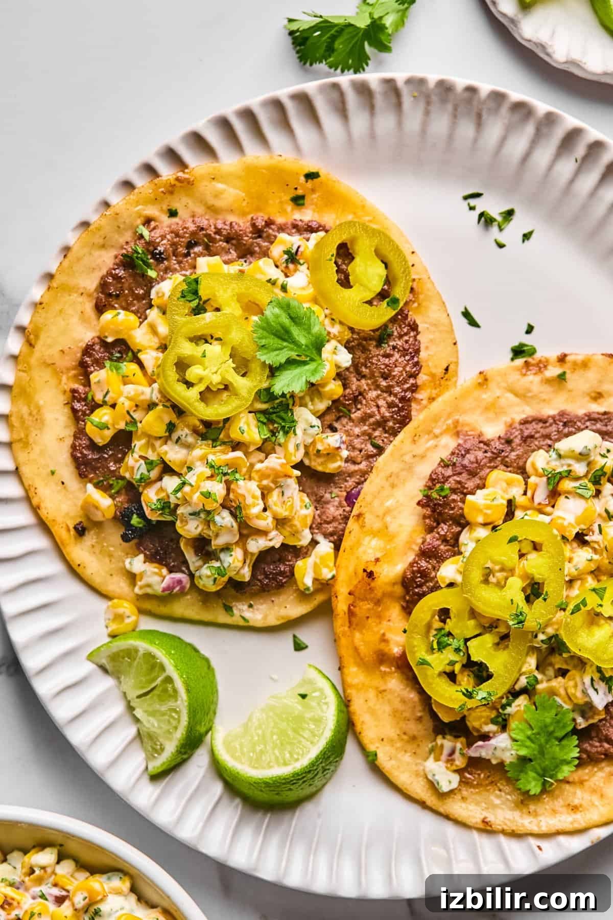 Street Corn Smash Tacos with Cotija and Lime 2 Two tacos on a white plate topped with ground meat, corn salad, pickled jalapeños, cilantro, and served with lime wedges.