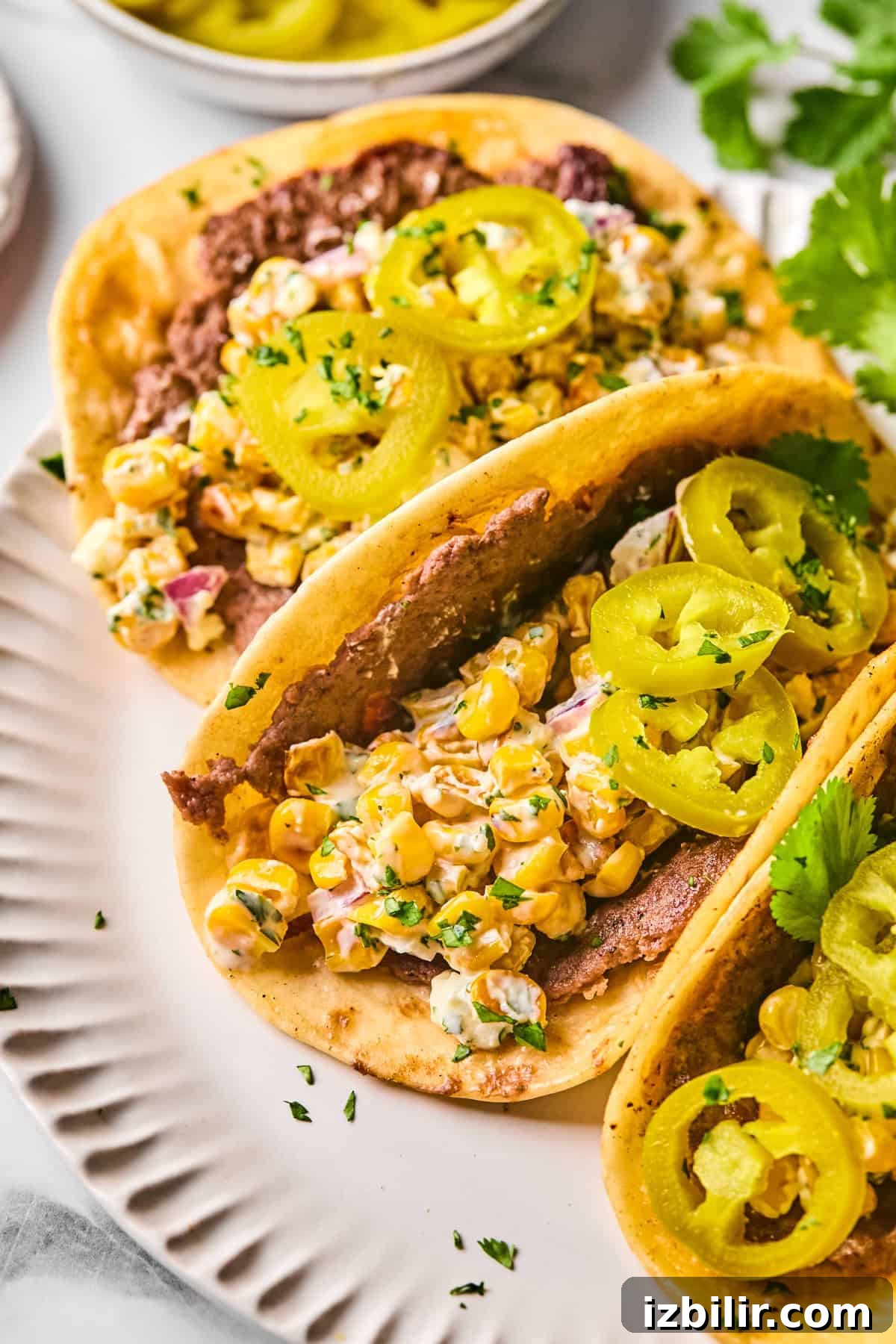 Street Corn Smash Tacos with Cotija and Lime 5 Two tacos on a white plate filled with refried beans, corn salad, and topped with sliced pickled jalapeños and chopped herbs.