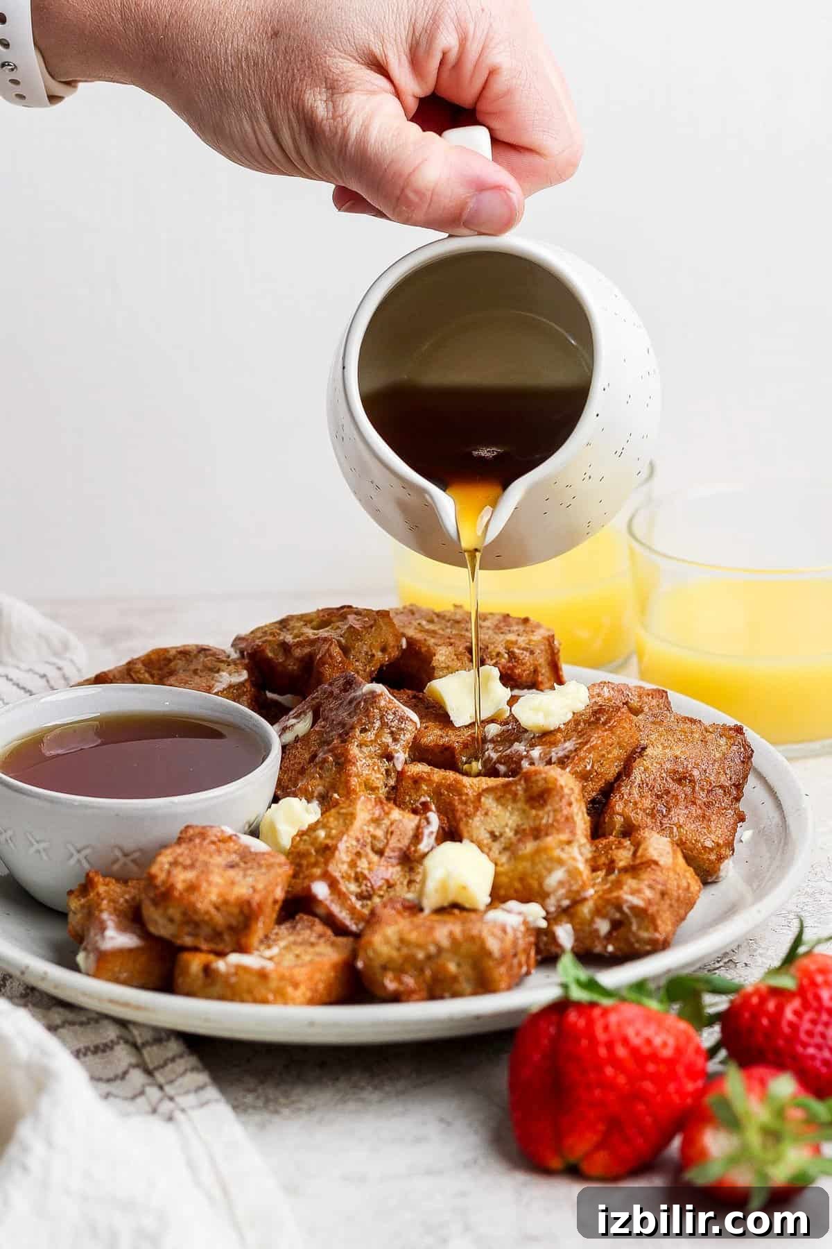 A hand pours syrup onto a plate of French toast cubes topped with butter. A bowl of syrup and strawberries are beside the plate. Two glasses of orange juice are in the background.