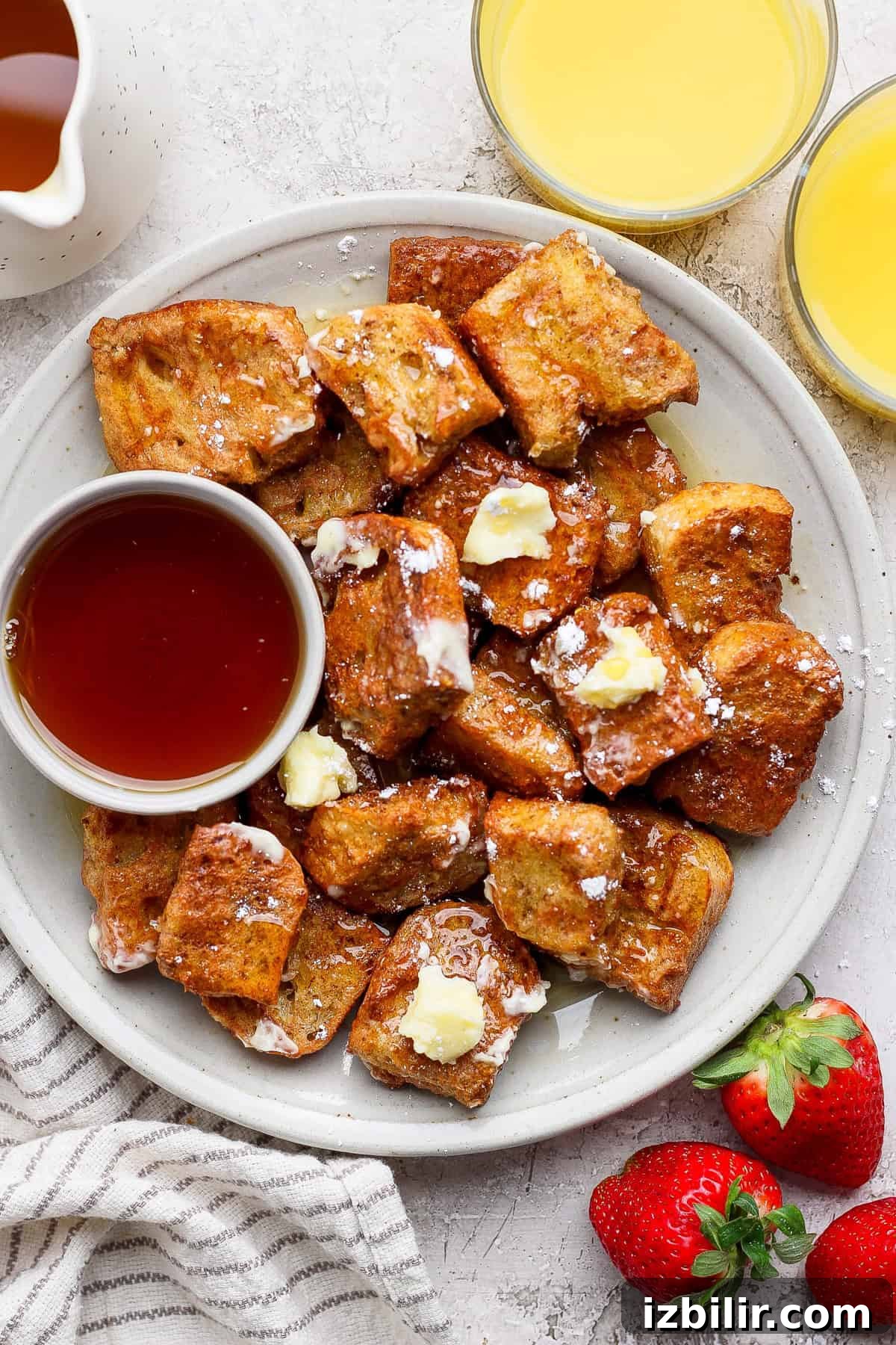 A plate of French toast bites with butter and powdered sugar, served with a small bowl of syrup. Two glasses of orange juice and fresh strawberries are on the side.