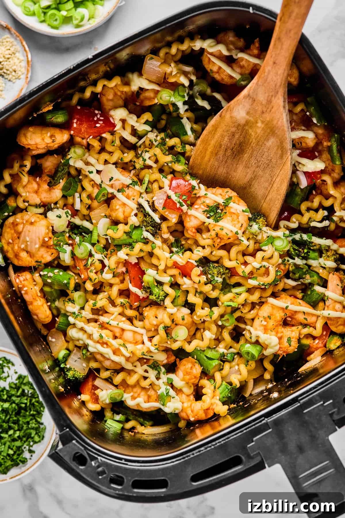 A wooden spoon stirs a mix of curly pasta, shrimp, vegetables, and herbs in an air fryer basket.