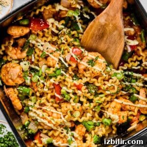 A pan filled with cooked pasta, shrimp, vegetables, and herbs being stirred with a wooden spatula.