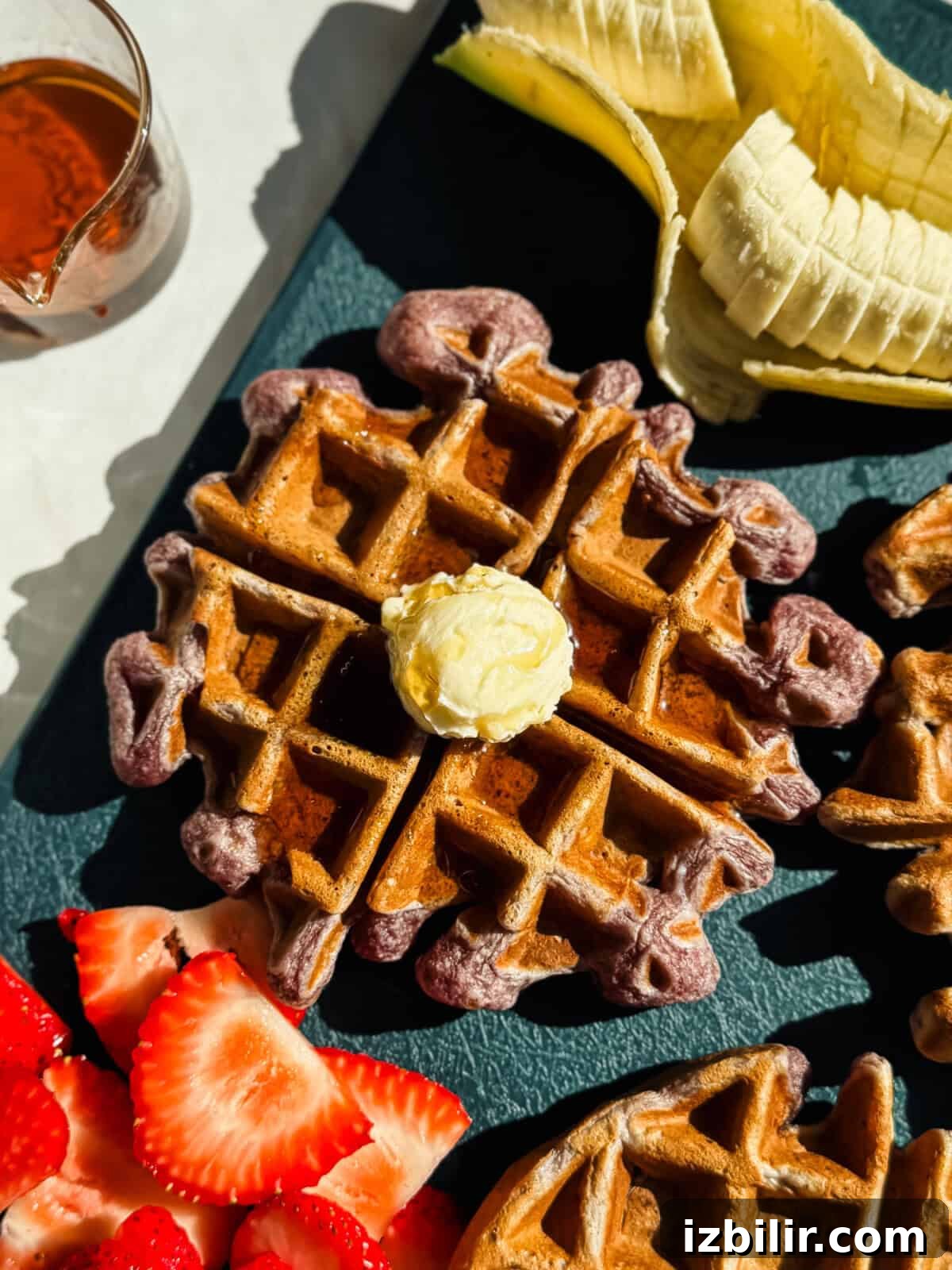 Strawberry Banana Cottage Cheese Waffles Recipe 2 A waffle topped with a pat of butter is shown alongside sliced bananas, strawberries, and a small glass of syrup on a dark surface.