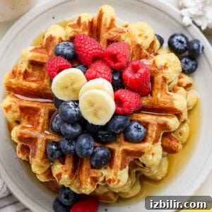 Strawberry Banana Cottage Cheese Waffles Recipe 3 A stack of waffles topped with blueberries, raspberries, banana slices, and drizzled with syrup on a round white plate.