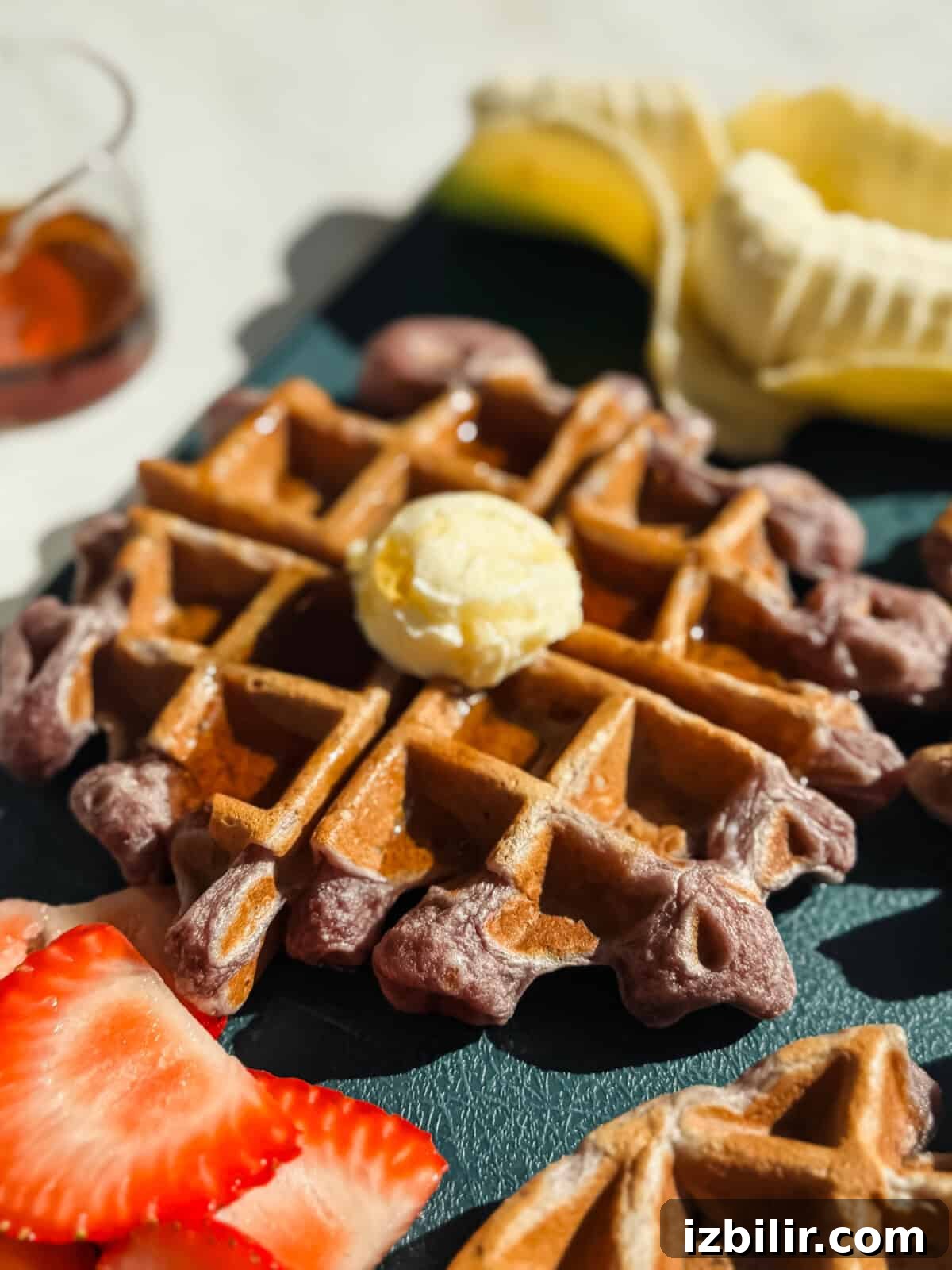 Strawberry Banana Cottage Cheese Waffles Recipe 4 A close-up of a waffle topped with a scoop of butter and syrup, with sliced strawberries and a partially peeled banana in the background.