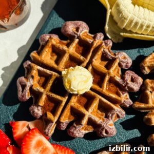 A waffle topped with a dollop of butter is surrounded by sliced bananas and strawberries on a dark surface.