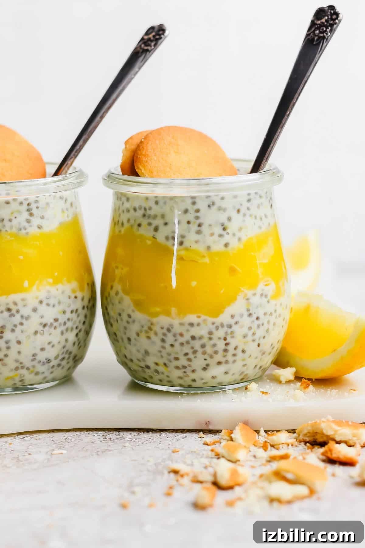 Silky Homemade Honey Lemon Curd for Toasts and Desserts 2 Two jars of chia pudding layered with lemon curd, each garnished with a cookie, placed on a marble surface with lemon wedges and cookie crumbs nearby.