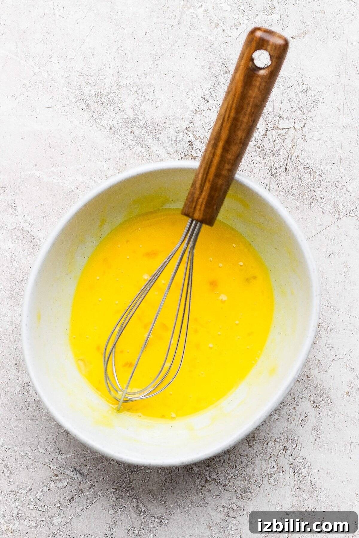Silky Homemade Honey Lemon Curd for Toasts and Desserts 3 Whisk in a bowl of beaten eggs on a textured, light-colored surface.