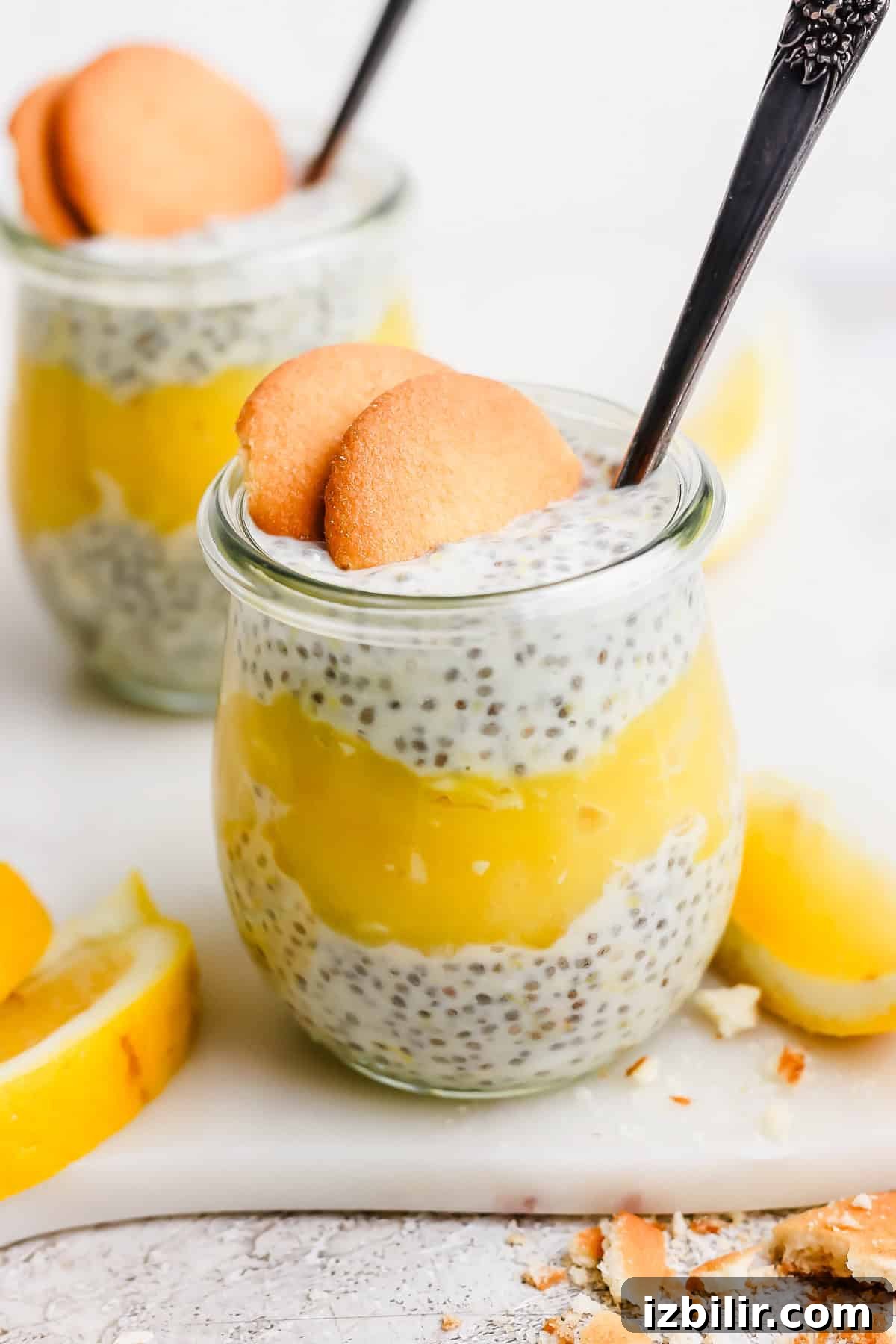 Two jars of chia pudding layered with yellow lemon curd, topped with two cookies and a spoon, surrounded by lemon slices.