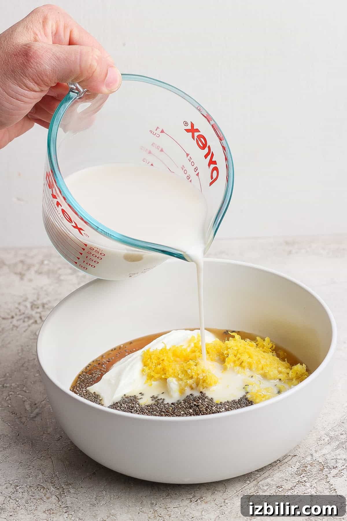 Pouring milk from a measuring cup into a bowl containing yogurt, chia seeds, lemon zest, and liquid.
