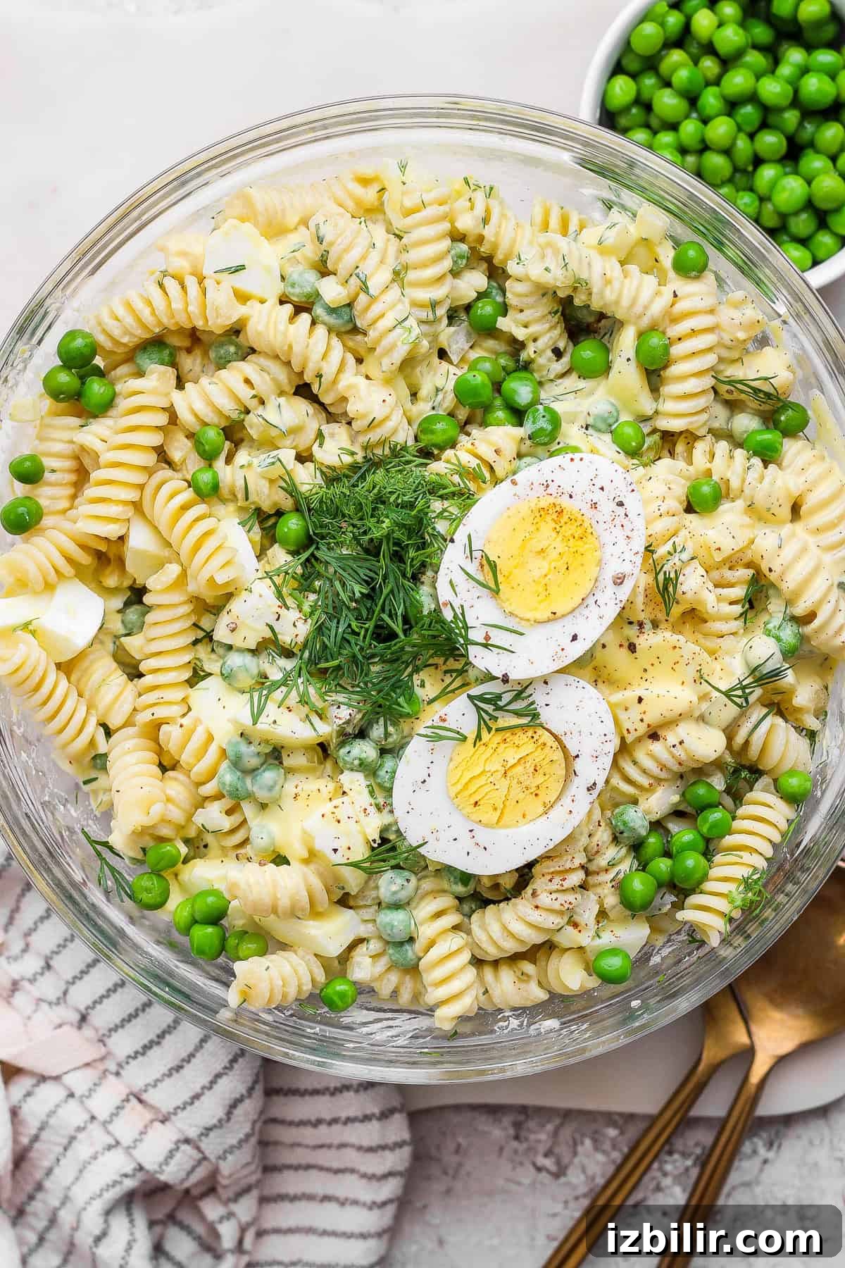 A bowl of pasta salad with rotini, green peas, hard-boiled egg slices, dill, and a creamy dressing. A gold spoon and a cloth napkin are next to the bowl.