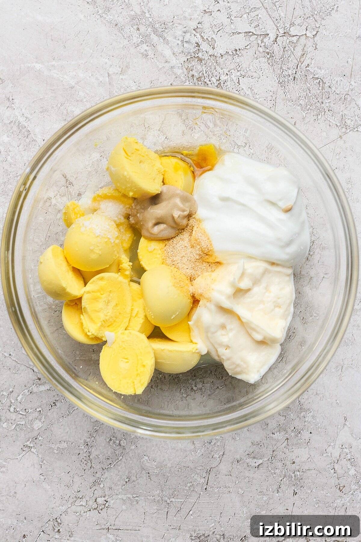 Bowl containing boiled egg yolks, mayonnaise, mustard, yogurt, and seasoning on a textured surface.