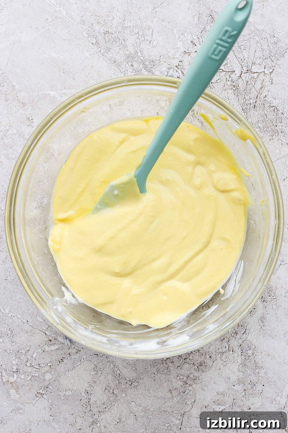 A glass bowl containing creamy, yellow batter with a teal spatula on a textured gray surface.