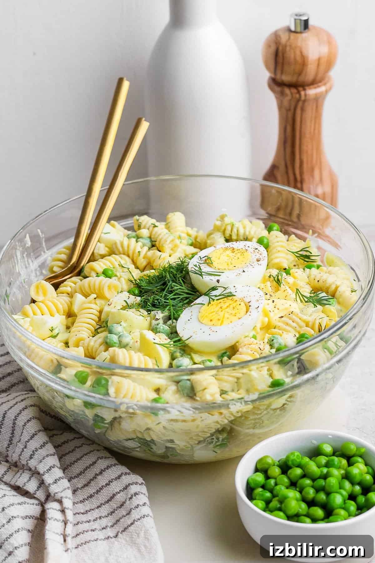 Glass bowl of pasta salad with sliced boiled eggs and herbs, garnished with peas. Placed on a table with chopsticks, a white towel, a small bowl of peas, a pepper mill, and an olive oil bottle.