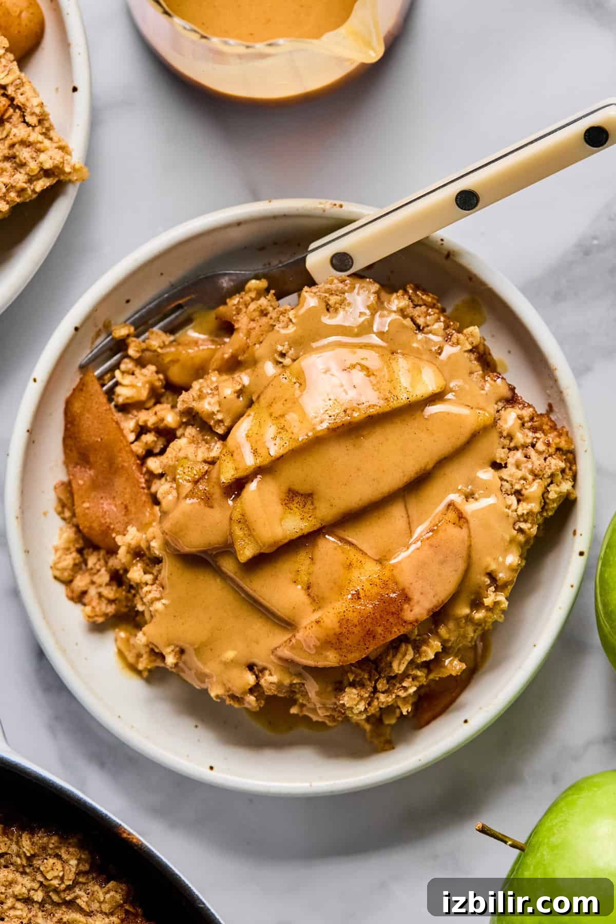 Baked Apple Oatmeal with Peanut Butter Caramel Sauce Recipe 2 A slice of apple crumble topped with sliced apples and a drizzle of caramel sauce on a white plate with a fork.
