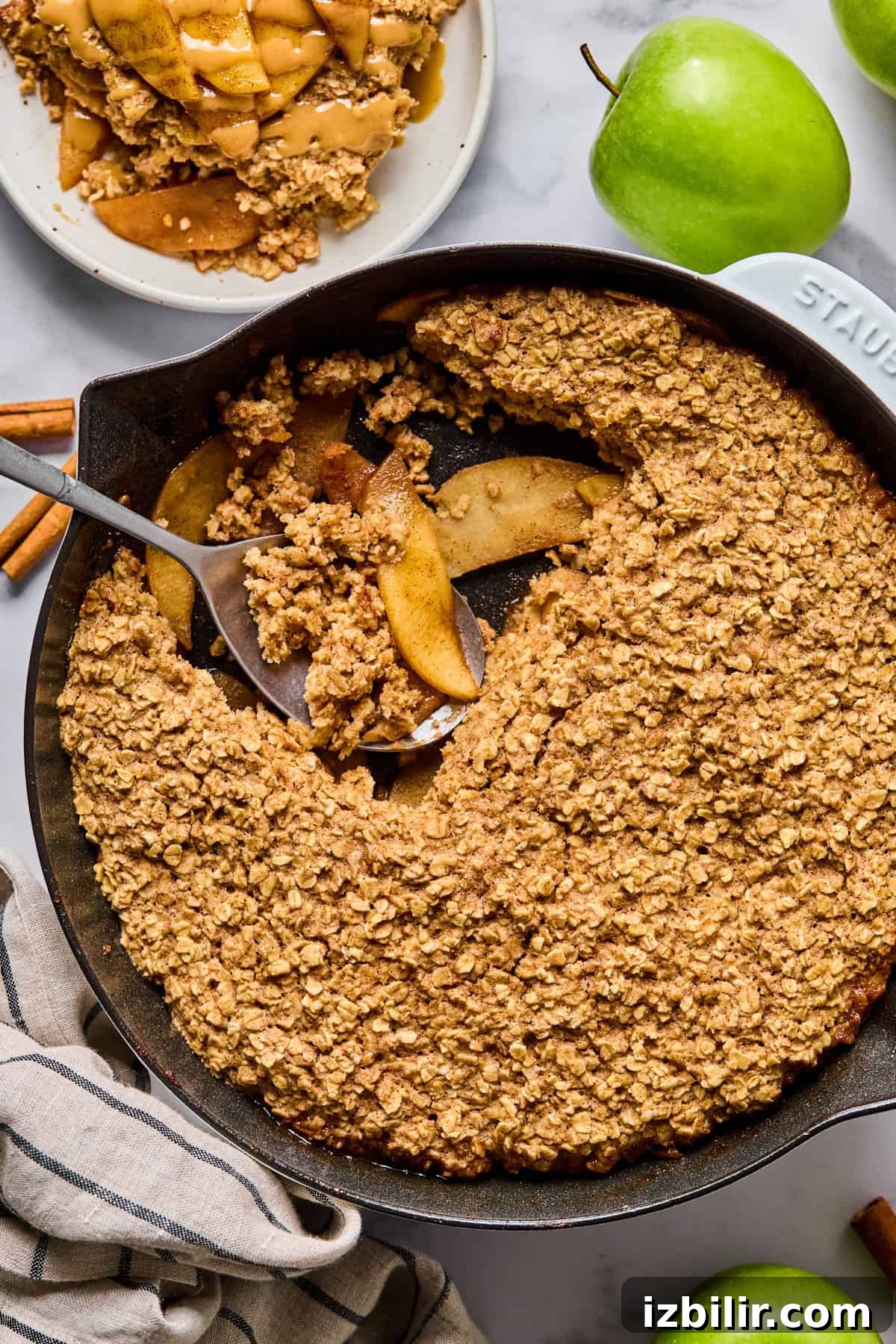Baked Apple Oatmeal with Peanut Butter Caramel Sauce Recipe 4 A cast iron skillet filled with baked apple crisp topped with oat crumble, with a serving scooped out onto a plate.