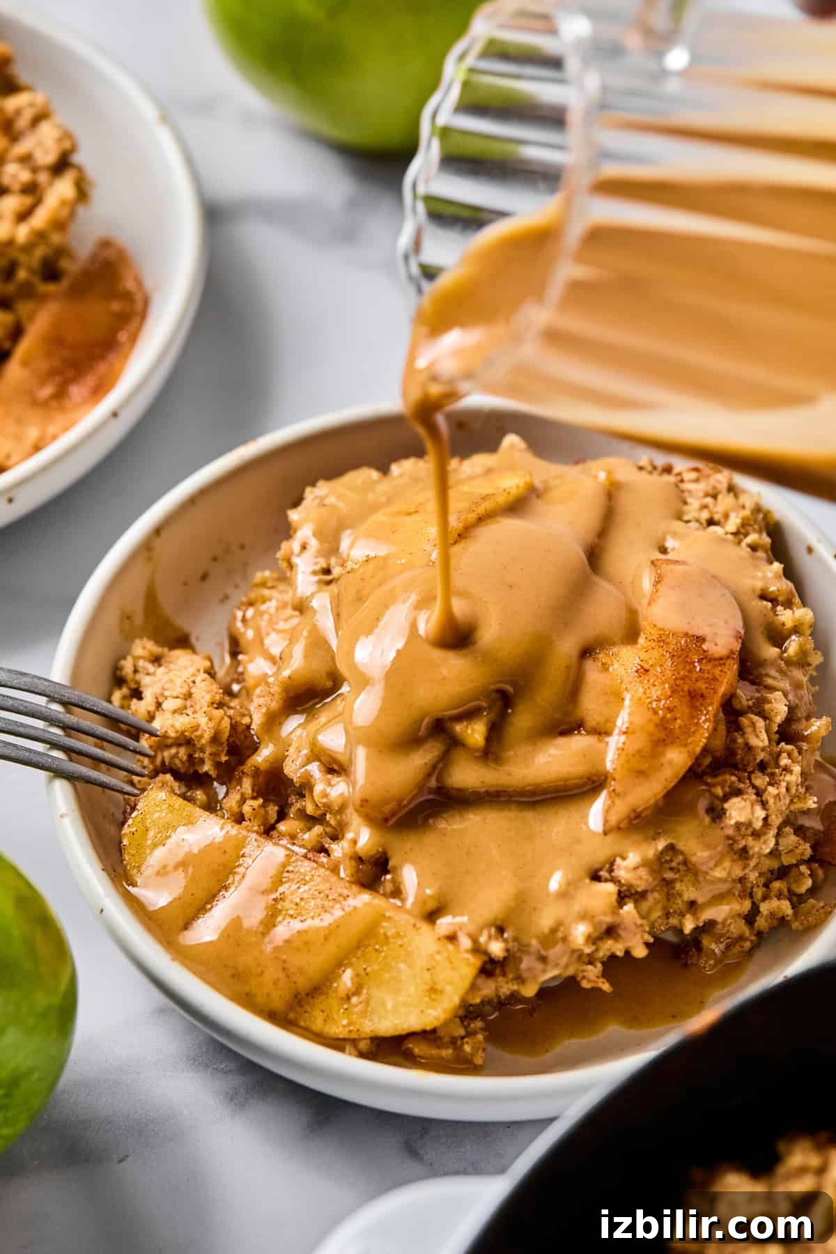 Baked Apple Oatmeal with Peanut Butter Caramel Sauce Recipe 5 A bowl of apple crisp topped with caramel sauce being poured from a glass container.